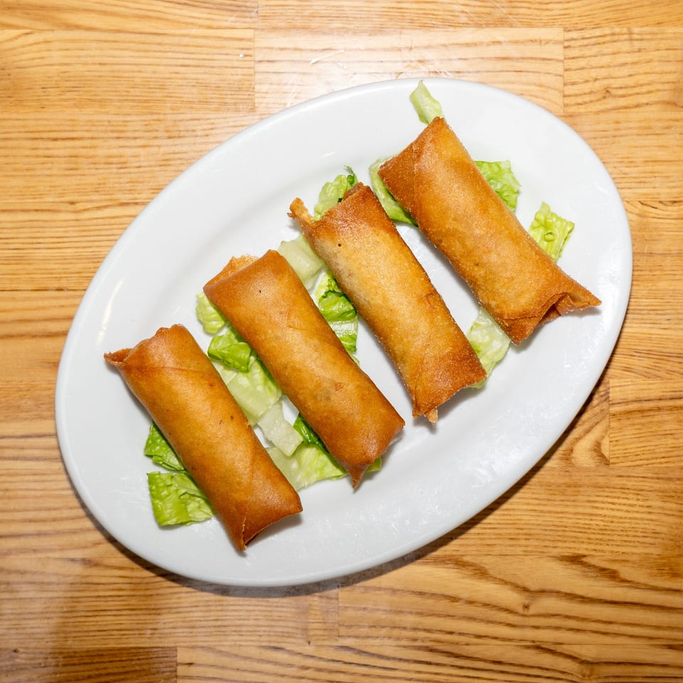 Four golden-brown fried spring rolls arranged on lettuce leaves atop a white plate on a wooden table.