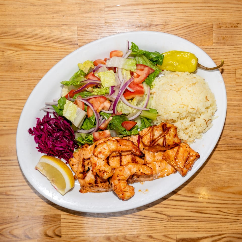 Grilled shrimp and salmon served with rice, garden salad, red cabbage, lemon, and pickled pepper on a white plate.