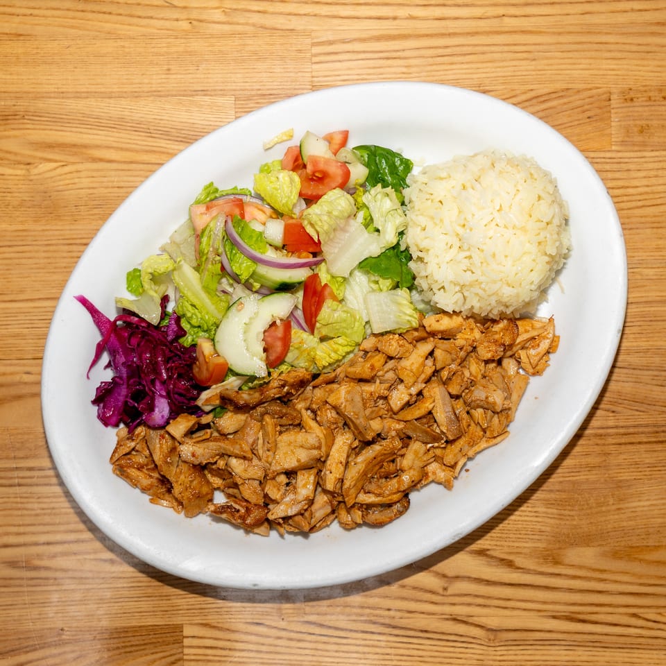 Plate of seasoned sliced chicken, white rice, and fresh lettuce, cucumber, tomato, onion, and cabbage salad on a wooden table.
