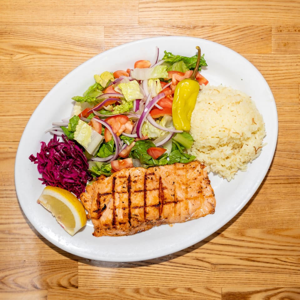 Grilled salmon served with rice, mixed salad, pickled cabbage, lemon wedges, and a yellow pepper on a white plate.