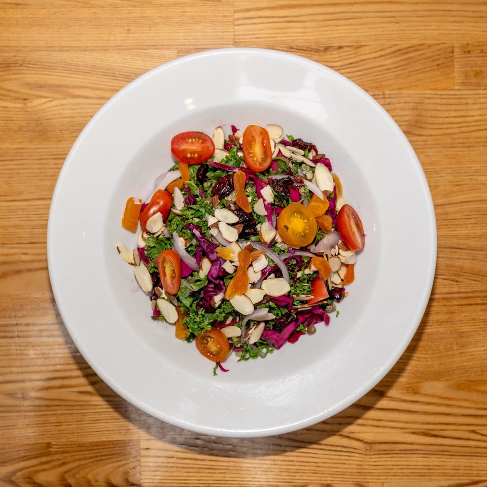 Colorful kale salad with tomatoes, red cabbage, almonds, dried cranberries, and apricots served on a white plate.