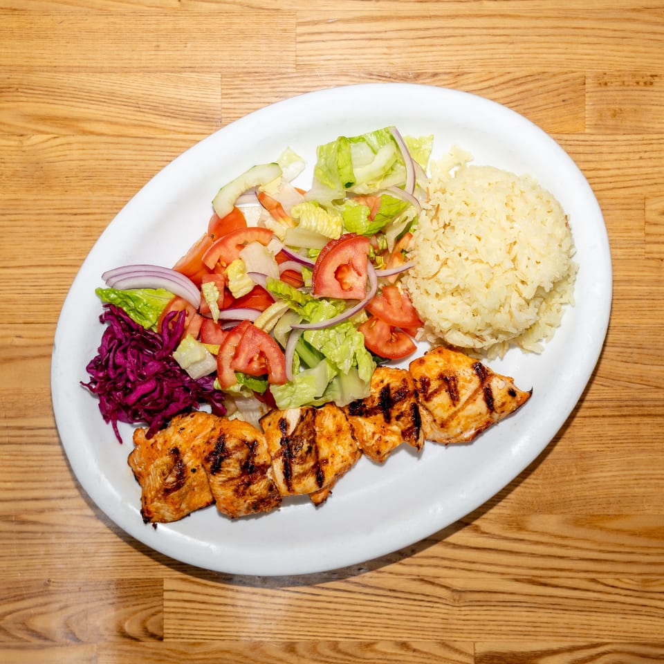 Grilled chicken served with rice, chopped salad, red cabbage, tomatoes, lettuce, onions, and cucumber on a white plate.