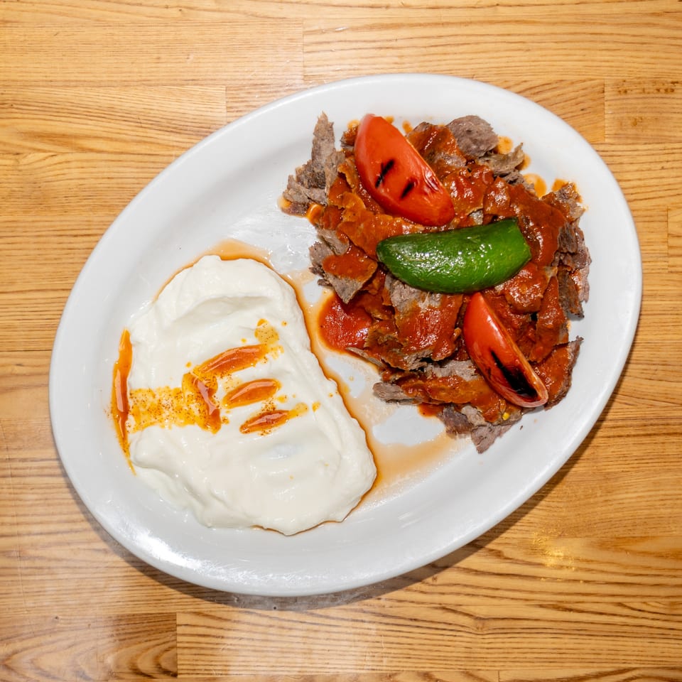 Plate of sliced meat topped with tomato, green pepper, and red sauce, served with creamy yogurt drizzled with chili oil.