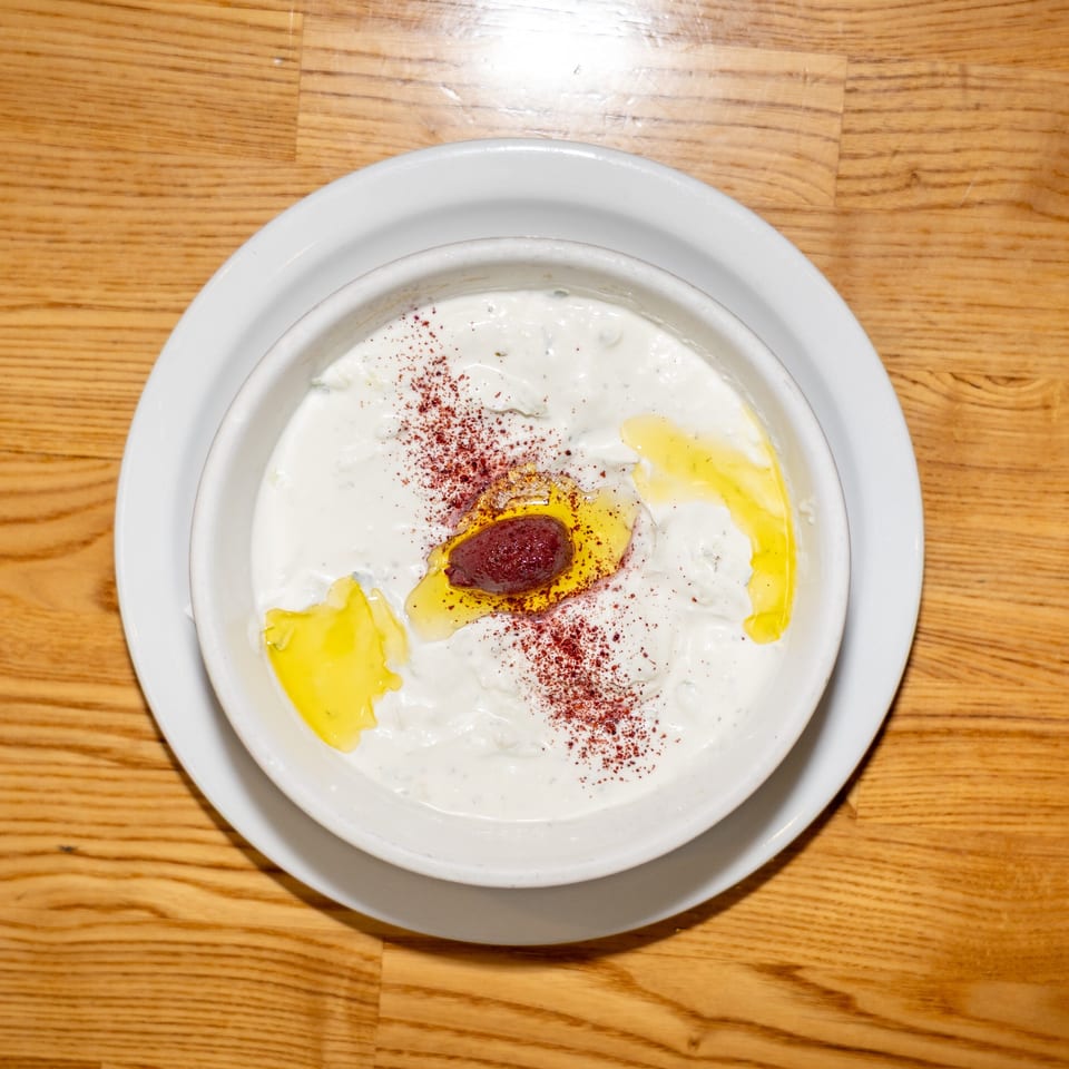 Bowl of creamy white dip topped with olive oil, red spice, and a dark olive, on a wooden table.