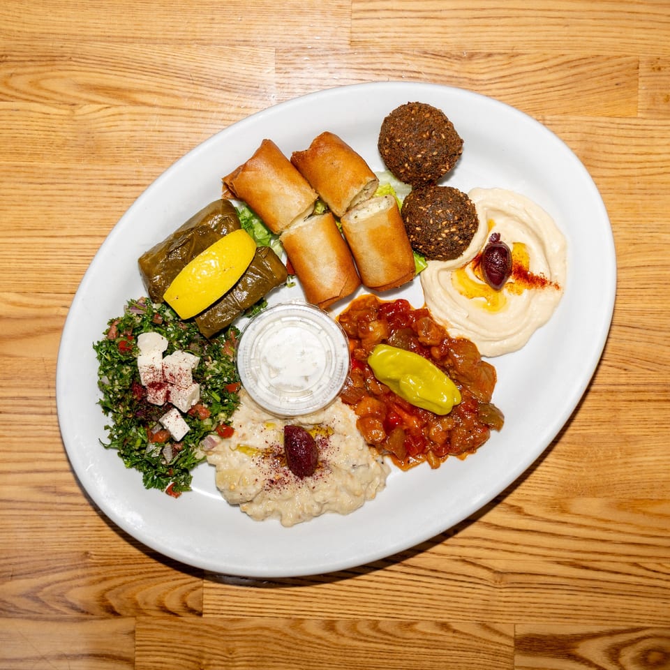 Mediterranean mezze platter with hummus, falafel, stuffed grape leaves, fried rolls, tabbouleh, and vegetables on a white plate.