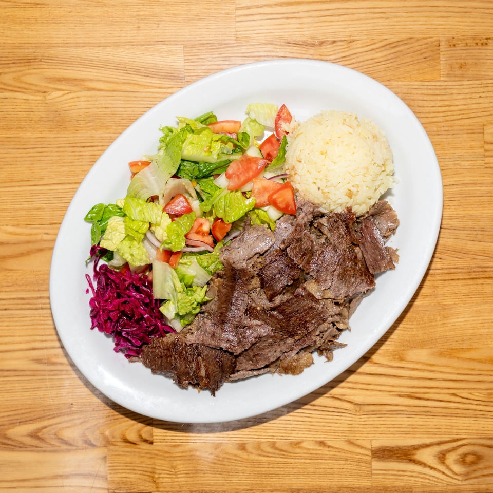 Plate of sliced grilled meat with yellow rice, lettuce and tomato salad, and shredded purple cabbage on a wooden table.