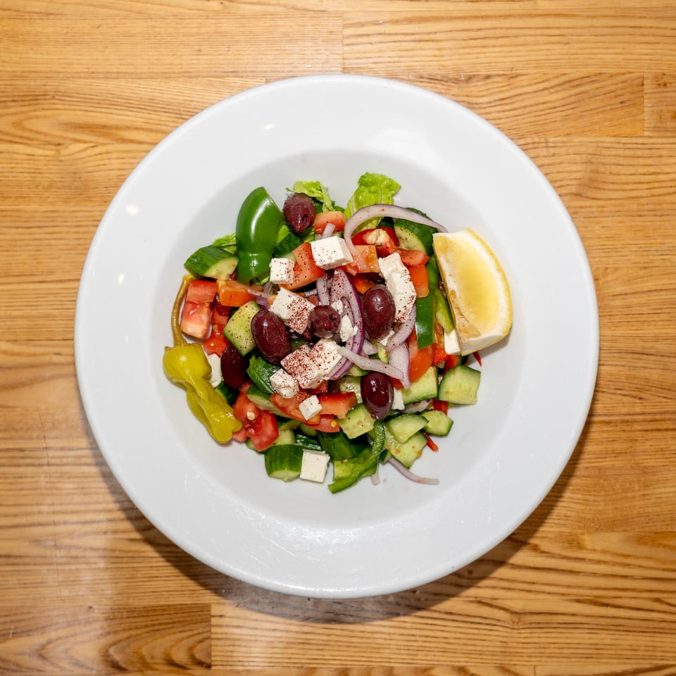 Greek salad with tomatoes, cucumbers, bell peppers, red onion, olives, feta cheese, pepperoncini, and lemon on a white plate.
