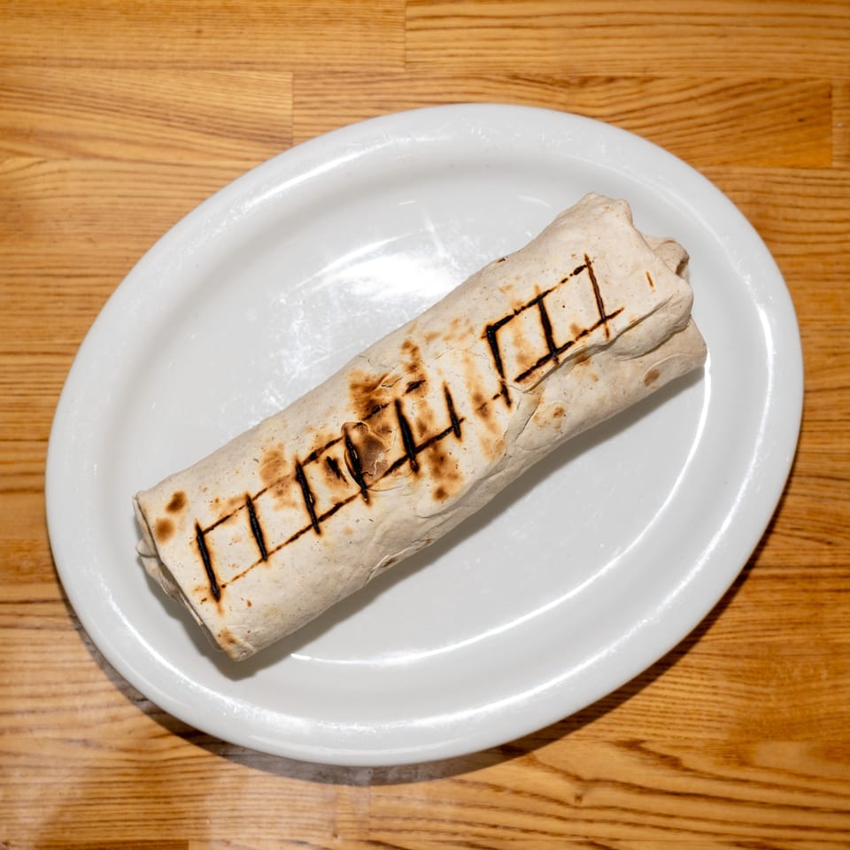Grilled burrito with dark grill marks served on a white plate atop a wooden restaurant table.