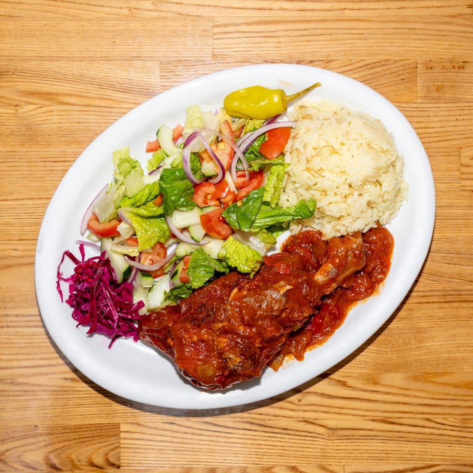 Plate of saucy grilled meat with rice, mixed salad, pickled cabbage, and a pepper on a wooden table.