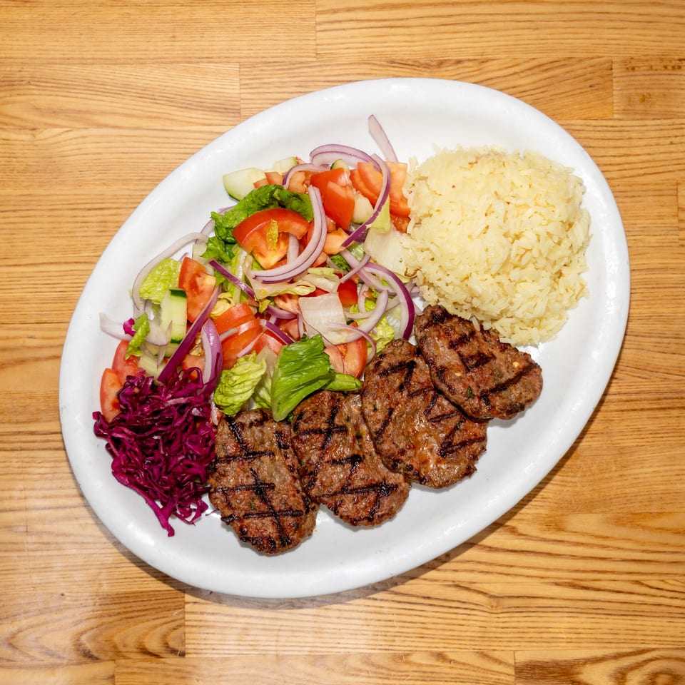 Grilled meat patties served with yellow rice, chopped salad, and shredded purple cabbage on a white plate.