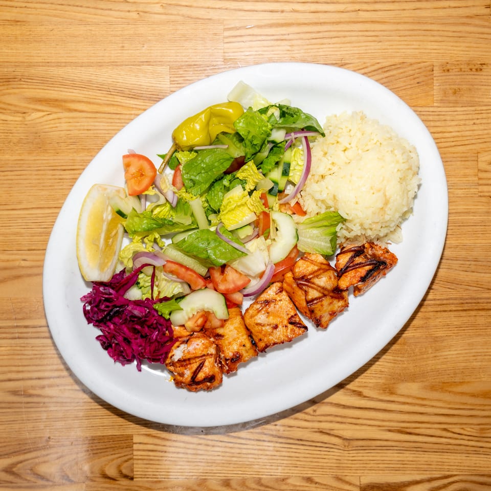 Plate of grilled chicken, white rice, mixed salad, pickled cabbage, lemon wedge, and peppers on a wooden table.