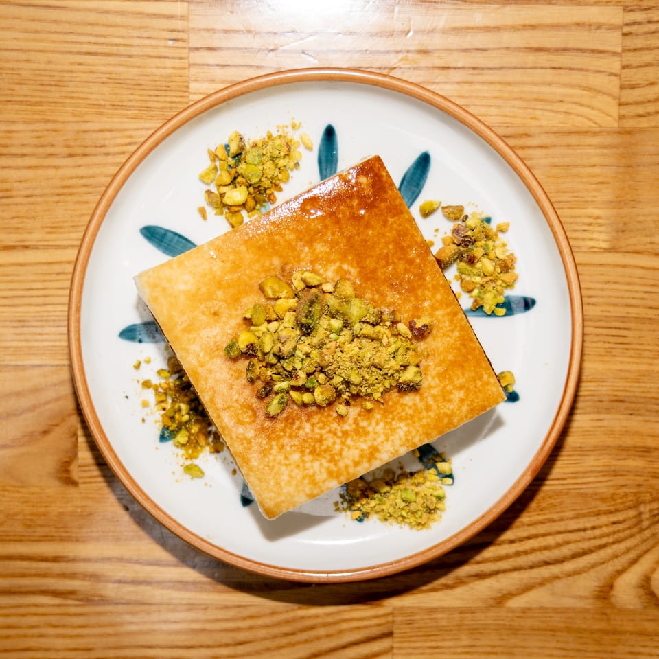 Golden rectangular pastry topped with crushed pistachios on a decorative plate over a wooden table.