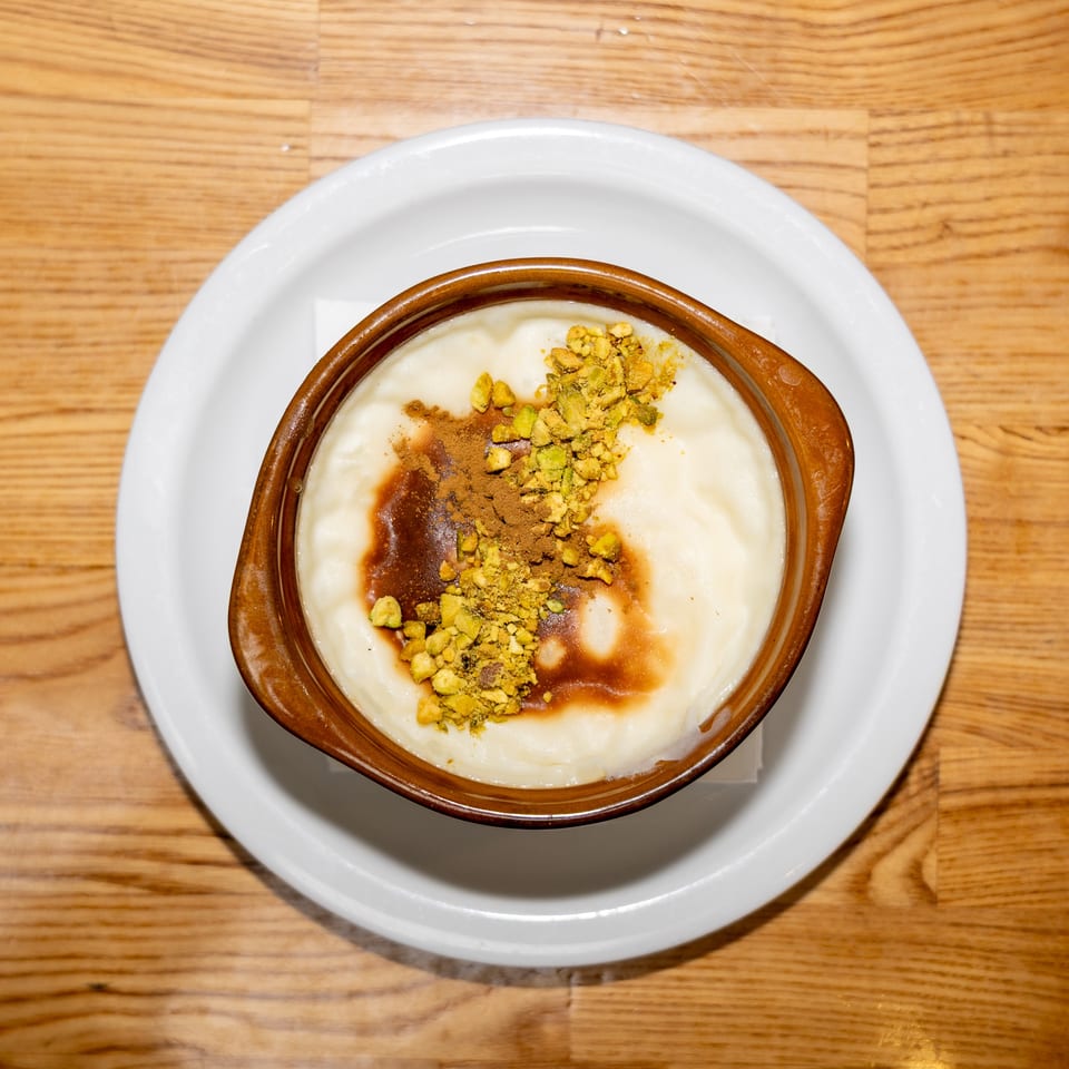 Creamy dessert topped with cinnamon, chocolate sauce, and crushed pistachios in a brown dish on a white plate.