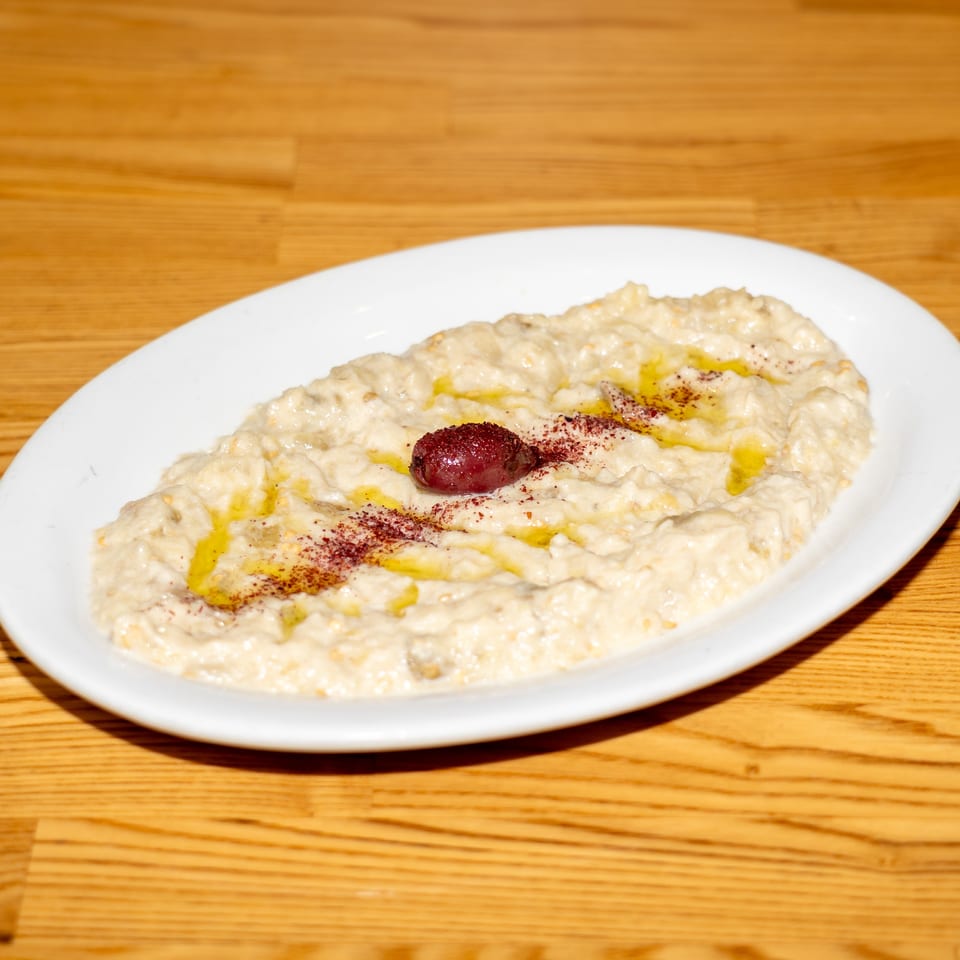Creamy hummus topped with olive oil, sumac, and a red garnish on a white plate atop a wooden table.