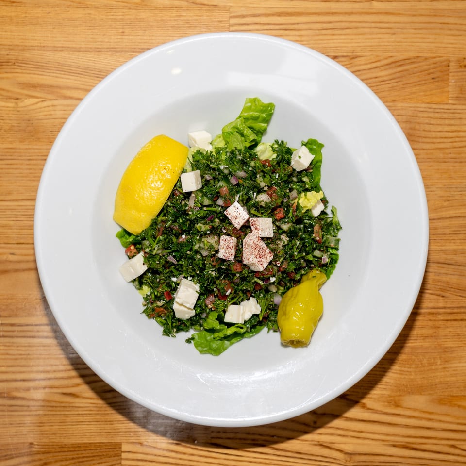 Tabbouleh salad with chopped herbs, vegetables, feta cheese, lemon wedge, and pickled pepper on a white plate.