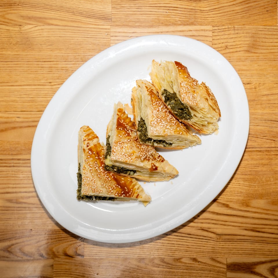 Sesame-topped flaky pastries filled with greens arranged on a white plate atop a wooden table.