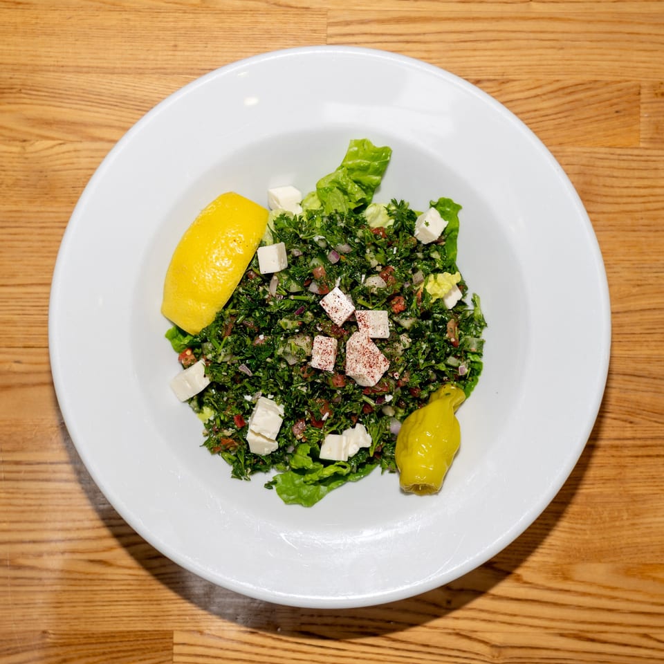 Tabbouleh salad with chopped parsley, tomatoes, onions, feta cheese, lettuce, lemon, and pickled pepper on a white plate.