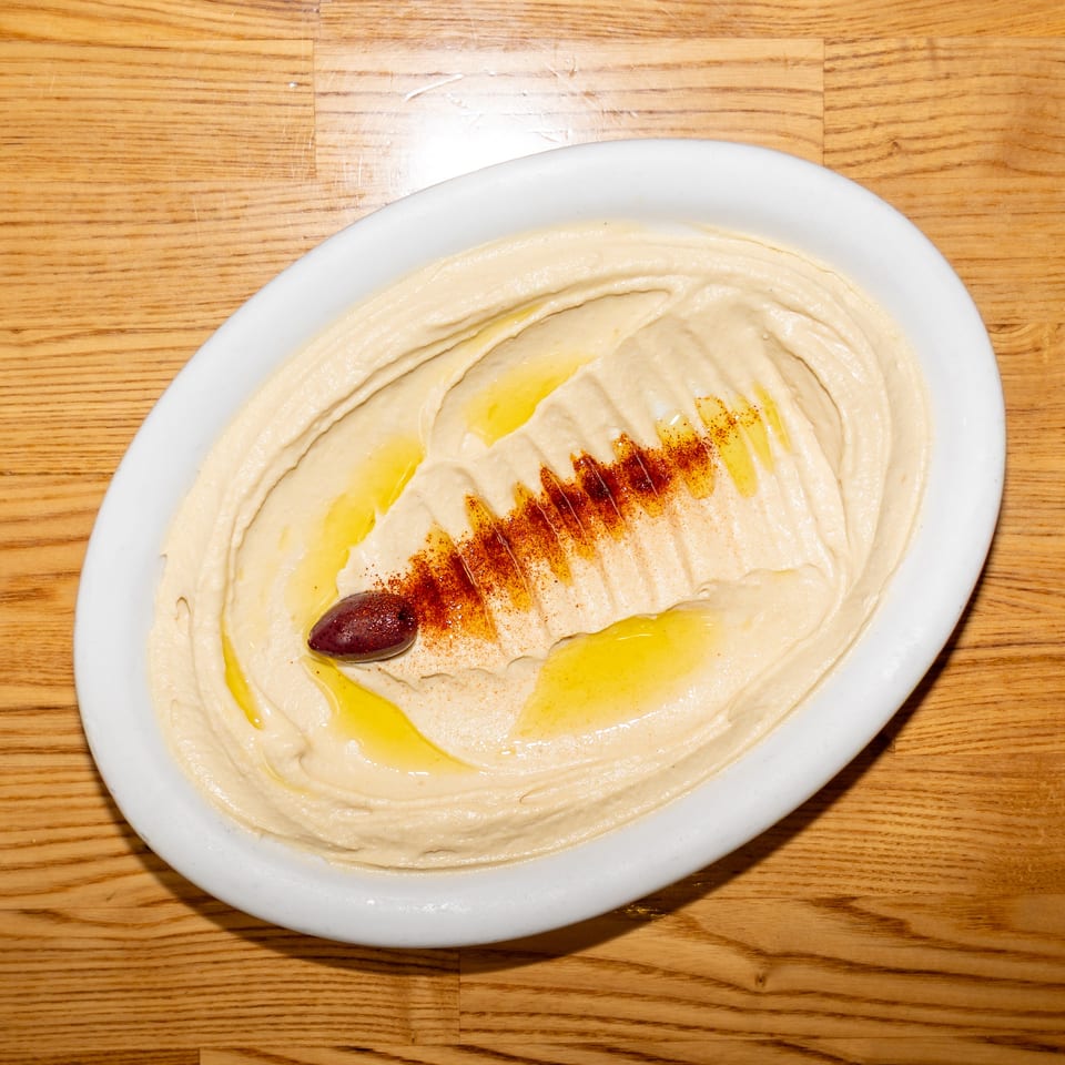 Creamy hummus topped with olive oil, paprika, and a kalamata olive on a white oval plate.
